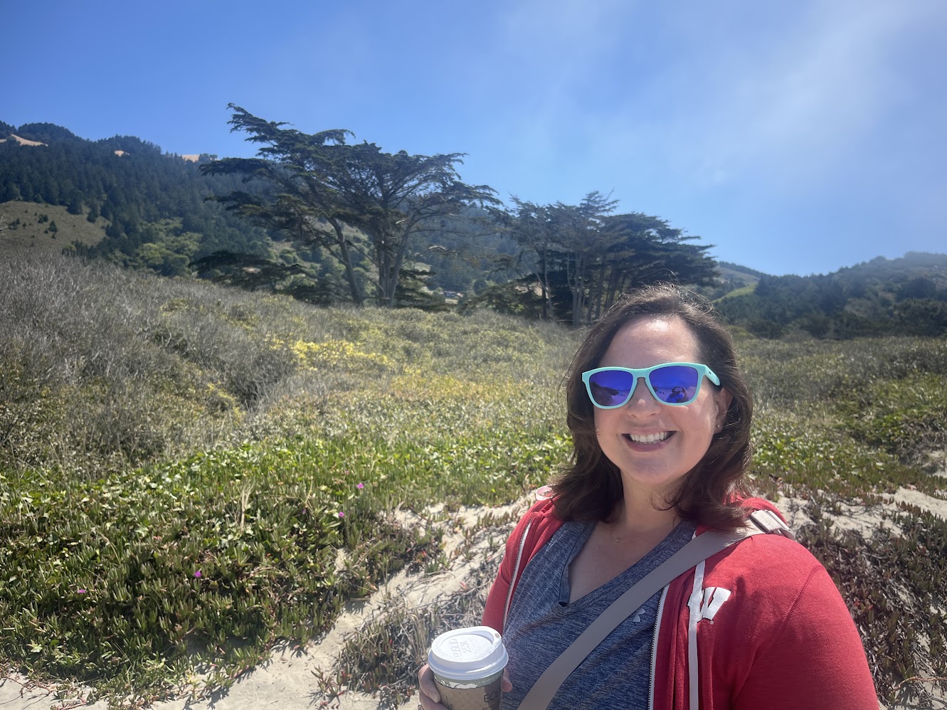 Amanda Heyman smiling on a sunny coastal Peninsula hike, holding a coffee with cypress-covered hills behind her