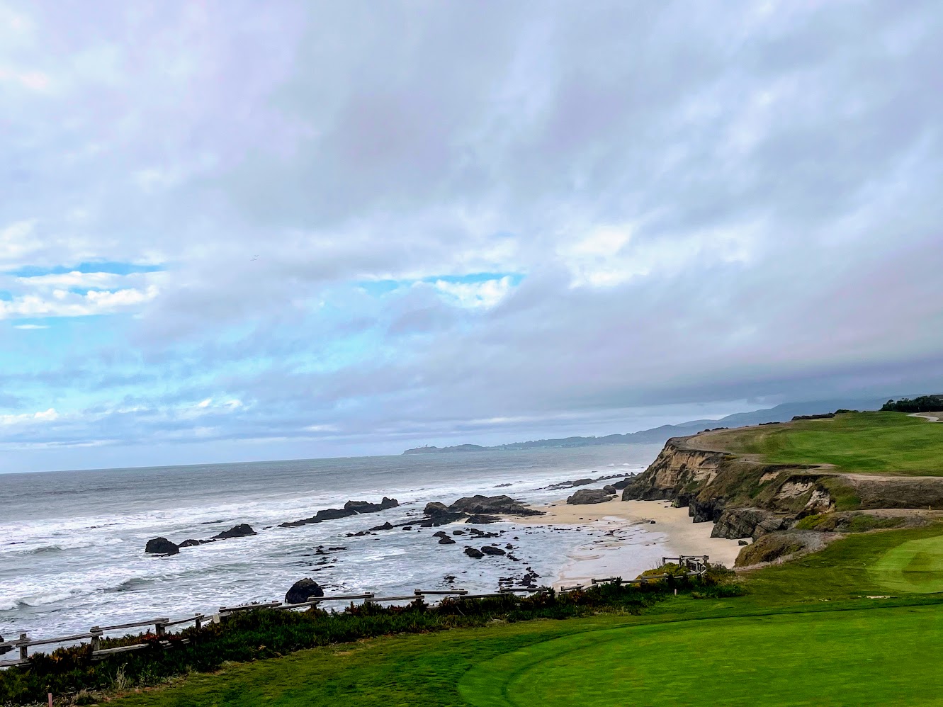 Ocean waves breaking over rocky reefs beneath the eroded bluffs and green bluff-top trail of the Half Moon Bay coast