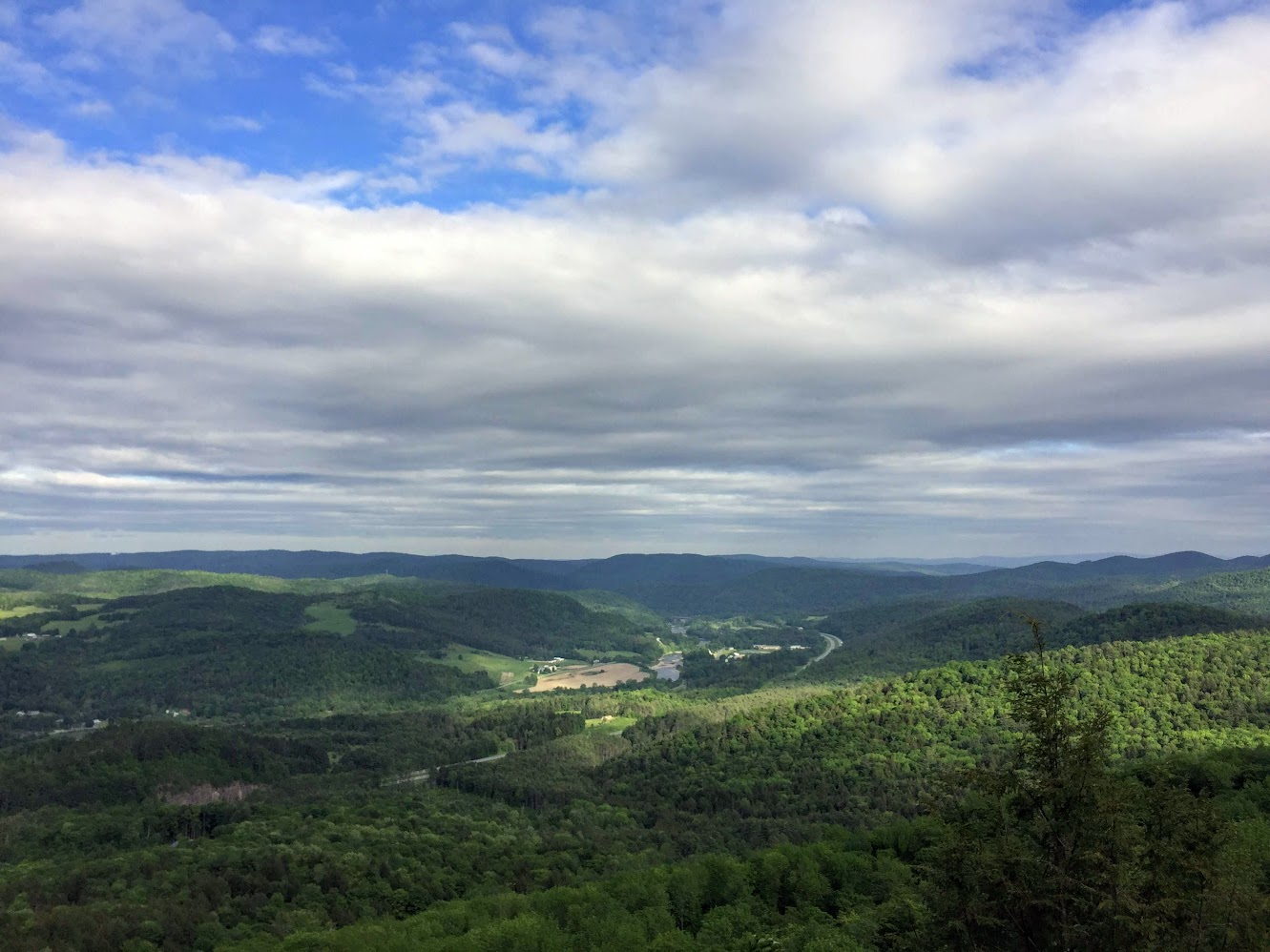 A sweeping hilltop panorama of rolling green forested hills fading into distant ridges under a wide cloudy sky